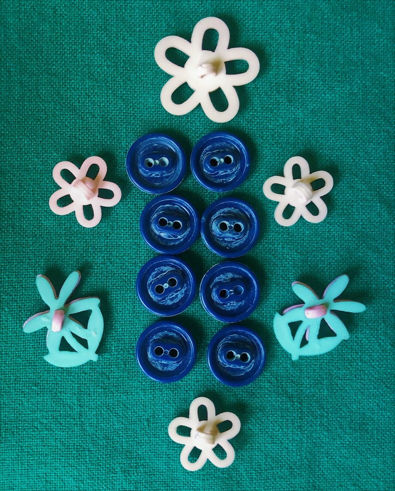 May include: An assortment of sewing buttons and embellishments on a green background. The collection includes blue round buttons, white and pink flower-shaped buttons, and turquoise butterfly-shaped embellishments.