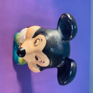 Pu&ograve; includere: Una statuetta di ceramica di Topolino, con orecchie nere, un naso nero e un viso bianco. La statuetta &egrave; dipinta con occhi marroni e una bocca gialla. La statuetta indossa un vestito blu e giallo.