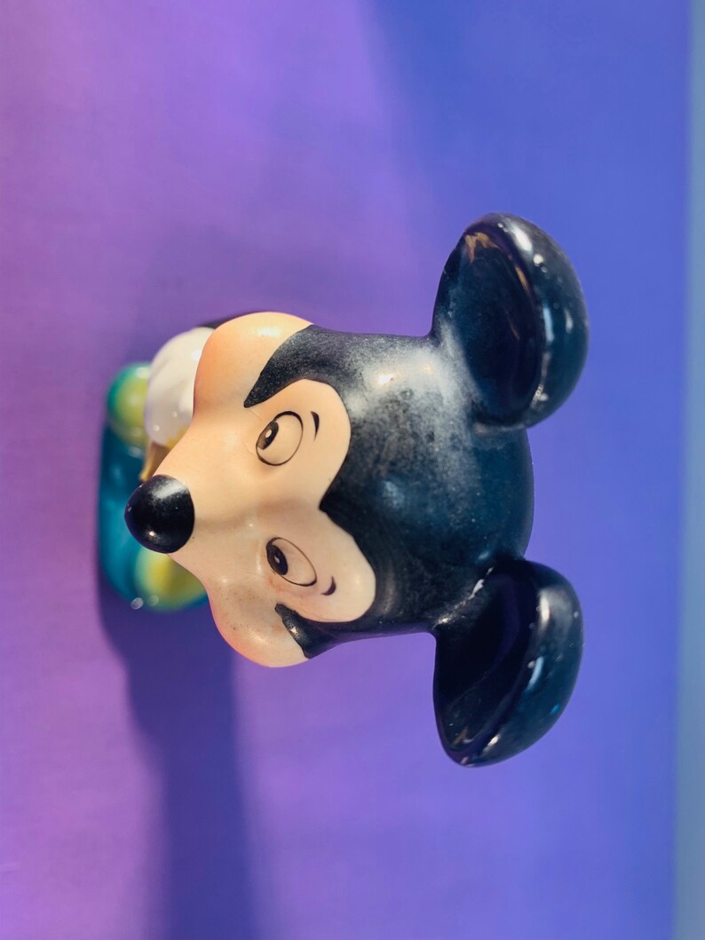 Pu&ograve; includere: Una statuetta di ceramica di Topolino, con orecchie nere, un naso nero e un viso bianco. La statuetta &egrave; dipinta con occhi marroni e una bocca gialla. La statuetta indossa un vestito blu e giallo.