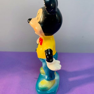 Pu&ograve; includere: Una statuetta in ceramica di Topolino in piedi su una base blu. Indossa una camicia gialla con un papillon rosso e pantaloni blu. Le sue orecchie sono nere e il suo viso &egrave; dipinto con occhi neri e un naso rosso.