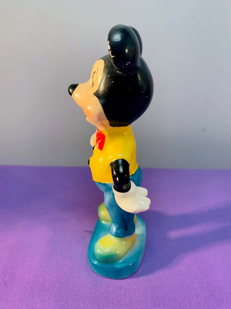 Pu&ograve; includere: Una statuetta in ceramica di Topolino in piedi su una base blu. Indossa una camicia gialla con un papillon rosso e pantaloni blu. Le sue orecchie sono nere e il suo viso &egrave; dipinto con occhi neri e un naso rosso.