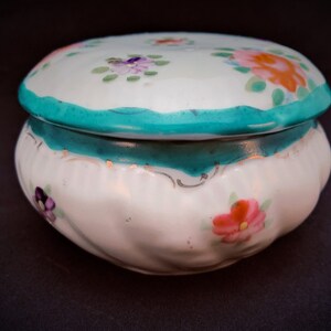 May include: A white ceramic trinket box with a lid, decorated with hand-painted flowers in pink, orange, and purple. The box has a teal scalloped edge and a matching teal band. The box is set against a dark background.
