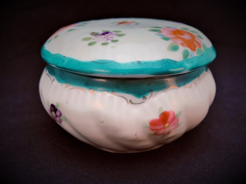 May include: A white ceramic trinket box with a lid, decorated with hand-painted flowers in pink, orange, and purple. The box has a teal scalloped edge and a matching teal band. The box is set against a dark background.