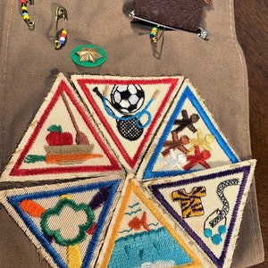 May include: Six triangular Girl Scout badges with colorful embroidered designs. The badges feature images of a soccer ball, a fish, a tree, a carrot, a gingerbread man, and a shirt.