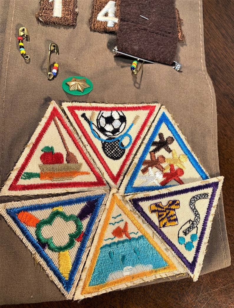 May include: Six triangular Girl Scout badges with colorful embroidered designs. The badges feature images of a soccer ball, a fish, a tree, a carrot, a gingerbread man, and a shirt.