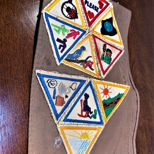 May include: A brown fabric sash with a collection of colorful triangular patches sewn on. The patches feature various images including a ladybug, a bird, a sun, and a person in a rocking chair. The text "PLEASE" is visible on one of the patches.