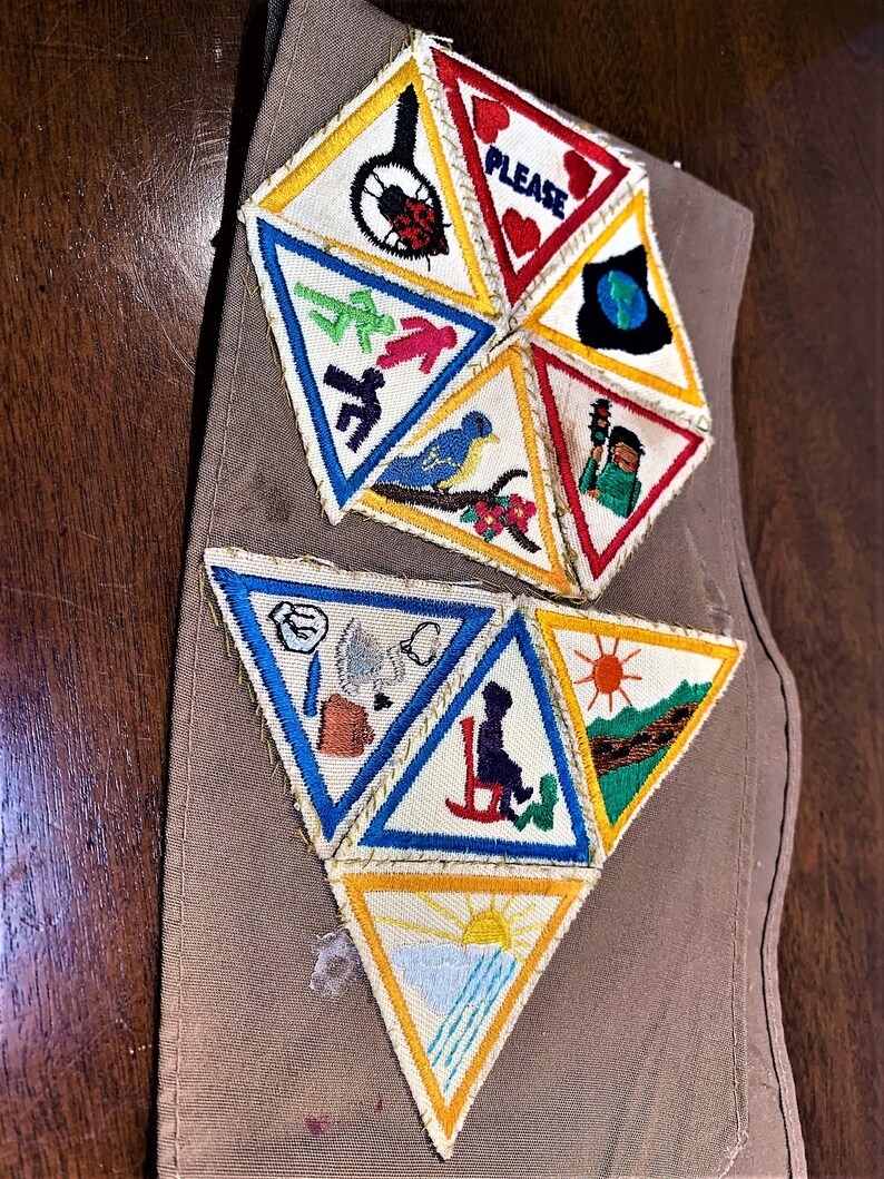 May include: A brown fabric sash with a collection of colorful triangular patches sewn on. The patches feature various images including a ladybug, a bird, a sun, and a person in a rocking chair. The text "PLEASE" is visible on one of the patches.