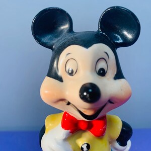 Pu&ograve; includere: Una statuetta in ceramica di Topolino che indossa una camicia gialla con un papillon rosso. Ha le orecchie nere e un naso nero. I suoi occhi sono spalancati e sta sorridendo.