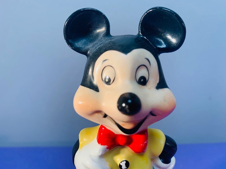 Pu&ograve; includere: Una statuetta in ceramica di Topolino che indossa una camicia gialla con un papillon rosso. Ha le orecchie nere e un naso nero. I suoi occhi sono spalancati e sta sorridendo.