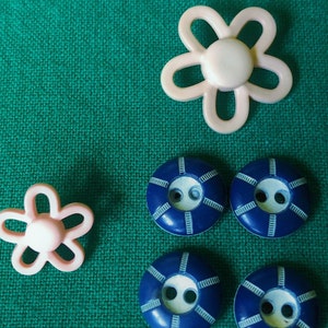 May include: An assortment of craft buttons in a variety of shapes and colors. Two light pink flower-shaped buttons and four navy blue buttons with white accents are displayed on a textured green fabric background. These buttons are ideal for sewing and crafting projects.