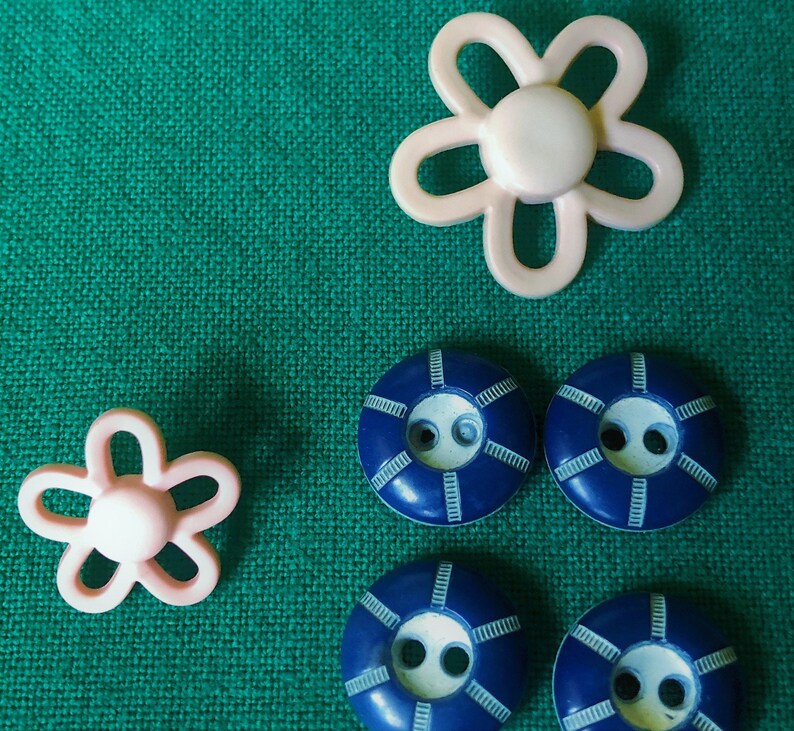 May include: An assortment of craft buttons in a variety of shapes and colors. Two light pink flower-shaped buttons and four navy blue buttons with white accents are displayed on a textured green fabric background. These buttons are ideal for sewing and crafting projects.