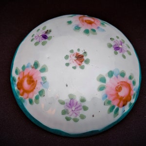 May include: A white, round ceramic dish with hand-painted floral designs. The flowers are pink, purple, and orange with green leaves. The rim is teal. The dish is likely for decorative or serving purposes.