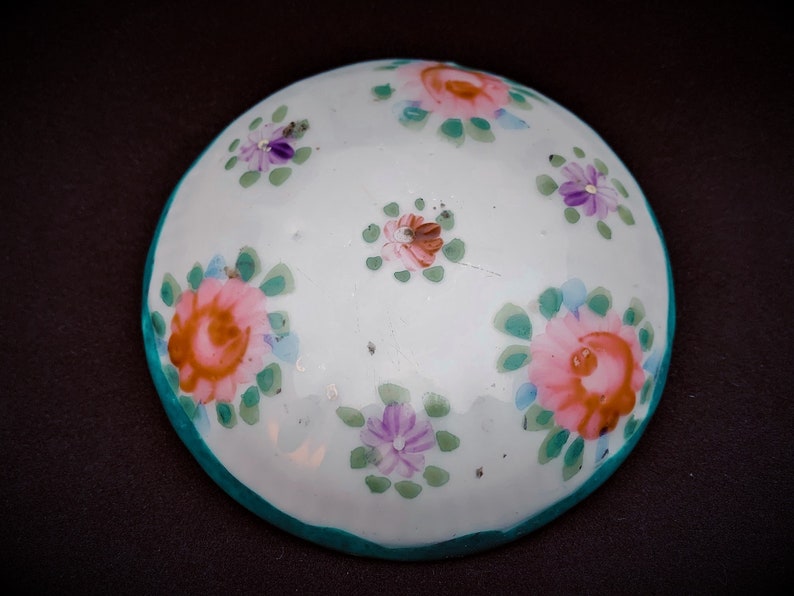 May include: A white, round ceramic dish with hand-painted floral designs. The flowers are pink, purple, and orange with green leaves. The rim is teal. The dish is likely for decorative or serving purposes.