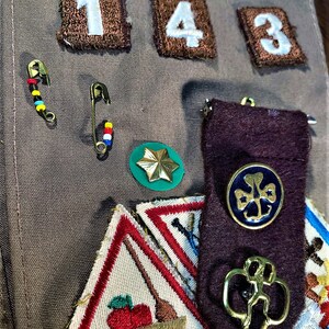 May include: A Girl Scout sash with various badges and patches, including a green four-leaf clover badge, a brown patch with the number "143", and a gold-colored pin with a four-leaf clover design.