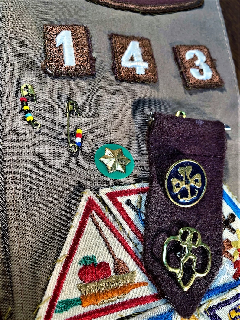 May include: A Girl Scout sash with various badges and patches, including a green four-leaf clover badge, a brown patch with the number "143", and a gold-colored pin with a four-leaf clover design.