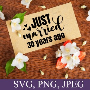 May include: A brown card with the text "Just married 30 years ago" in black lettering with three small hearts. The card is surrounded by white flowers on a brown wooden surface.