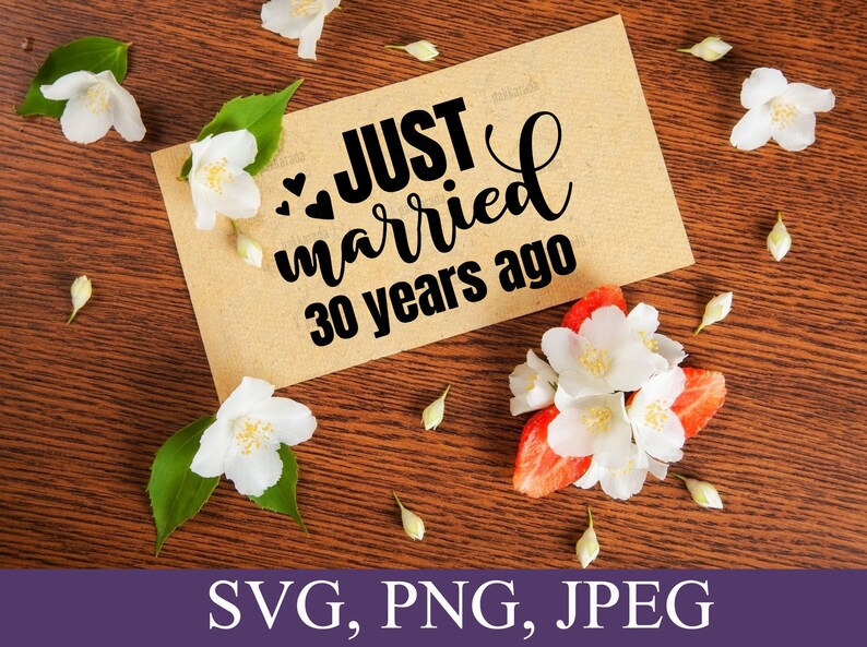 May include: A brown card with the text "Just married 30 years ago" in black lettering with three small hearts. The card is surrounded by white flowers on a brown wooden surface.