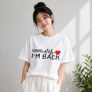May include: White t-shirt with the text "ANNNND... I'M BACK" in black, a red heart, and a heartbeat line design. The shirt is a casual, short-sleeved style.