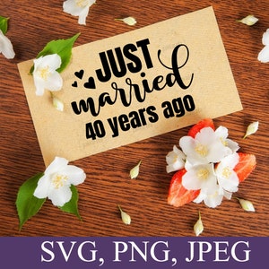 May include: A brown card with the text "JUST married 40 years ago" in black script font. The card is surrounded by white flowers on a brown wooden surface.