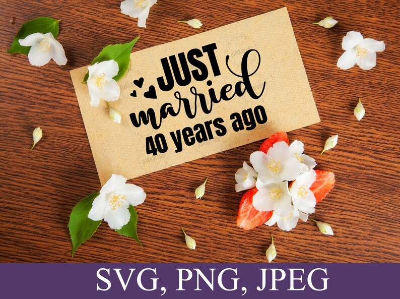 May include: A brown card with the text "JUST married 40 years ago" in black script font. The card is surrounded by white flowers on a brown wooden surface.