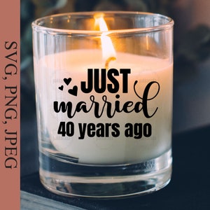 May include: A clear glass candle jar with a white candle inside. The candle has a black inscription that reads "JUST married 40 years ago" with two small hearts.
