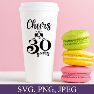 May include: A white travel coffee cup with a lid, featuring the text "Cheers to 30 years" in black script and a graphic of clinking champagne glasses. Three colorful macarons in green, yellow, and pink are stacked beside the cup.