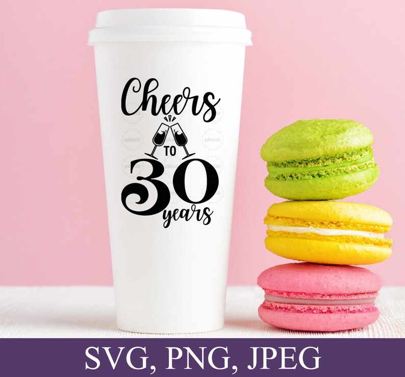 May include: A white travel coffee cup with a lid, featuring the text "Cheers to 30 years" in black script and a graphic of clinking champagne glasses. Three colorful macarons in green, yellow, and pink are stacked beside the cup.