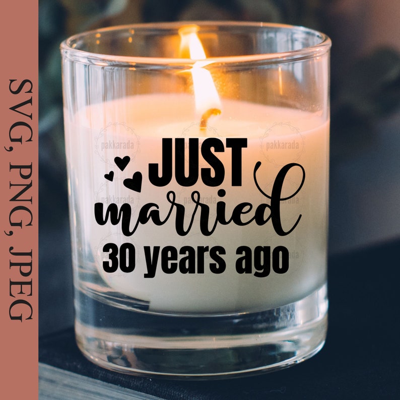May include: A clear glass candle jar with a white candle inside. The candle has a black inscription that reads "JUST married 30 years ago" with two small hearts above the word "married".