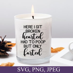 May include: A white candle with a black inscription that reads "Here I sit broken hearted had to poop but only farted". The candle is lit and sitting on a table with fall foliage.