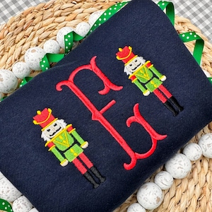 May include: Navy blue sweatshirt with a red embroidered letter "F" and two embroidered nutcracker figures in green, red, and gold. The sweatshirt is surrounded by white beaded garland and green ribbon.