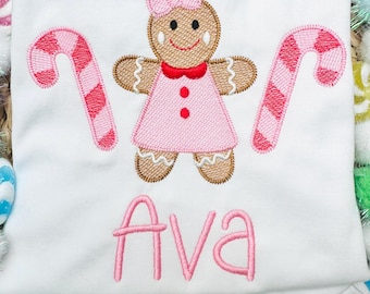 May include: White shirt with a pink gingerbread girl and candy cane design. The gingerbread girl wears a pink dress and bow. The name "Ava" is embroidered in pink script below the design. The shirt has a ruffled hem.