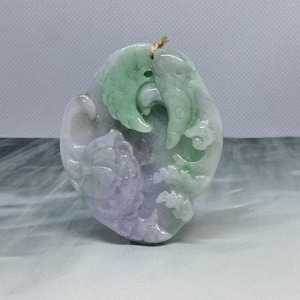 May include: An oval-shaped carved jade pendant featuring a light green and lavender color palette. The stone is intricately sculpted with a bird-like figure at the top and a floral or fruit-like cluster at the bottom, with a small string loop.