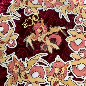 May include: A selection of stickers and a keyring featuring a cartoon pony with a distinctive design. The pony has a tan body, pink mane and tail, and red eyes. The stickers and keyring have a gold-coloured outline and a gold-coloured keyring clasp.