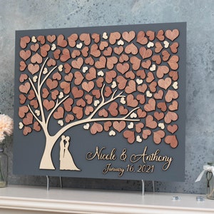 May include: A grey wooden guest book with a tree design. The tree is made of wooden hearts in shades of brown and beige. The text "Nicole & Anthony January 16, 2021" is written on the bottom of the guest book.