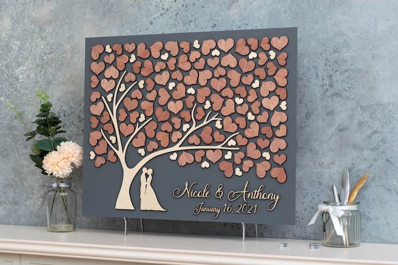 May include: A grey wooden guest book with a tree design. The tree is made of wooden hearts in shades of brown and beige. The text "Nicole & Anthony January 16, 2021" is written on the bottom of the guest book.