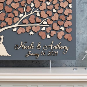 May include: A wooden heart tree guest book with a grey background. The tree is decorated with wooden hearts in shades of brown and white. The names "Nicole & Anthony" and the date "January 16, 2021" are written in cursive on the sign.