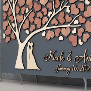 May include: A grey wooden guest book with a tree design and a couple silhouette. The tree is decorated with wooden hearts. The text "Nicole & Anthony January 16, 2021" is written on the bottom of the guest book.