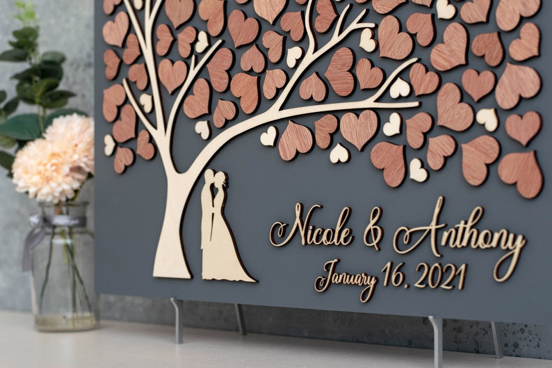 May include: A grey wooden guest book with a tree design and a couple silhouette. The tree is decorated with wooden hearts. The text "Nicole & Anthony January 16, 2021" is written on the bottom of the guest book.