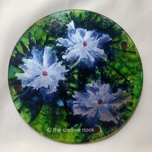 May include: A round glass coaster with a green, blue, and white floral design. The coaster has a textured surface and features three white flowers with red centers. The text "© the cre8ive nook" is printed on the bottom of the coaster.