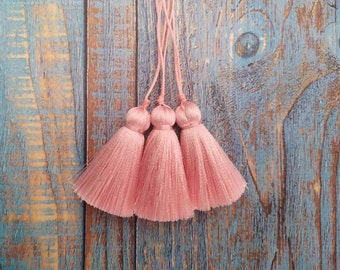May include: Three pink tassel trims with a light pink color. The tassels are hanging from a wooden surface.