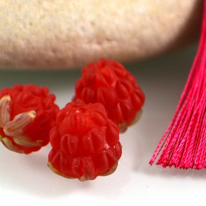 May include: Three red glass beads shaped like raspberries. The beads have a glossy finish and are arranged on a white surface.