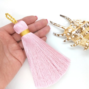 May include: A pink tassel with a gold cord and a gold trim. The tassel is approximately 12.5 cm long and is made of soft, silky yarn.