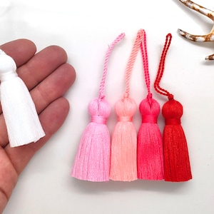 May include: Five colorful tassels in shades of pink, red, and white. The tassels are made of soft yarn and have a delicate fringe.