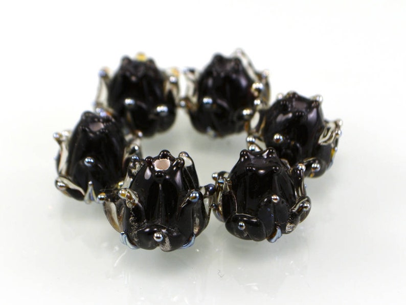 May include: A circular arrangement of six glossy black frog-shaped beads. Each bead has silver-colored accents and small, iridescent details. The beads are arranged on a white surface, creating a decorative pattern.
