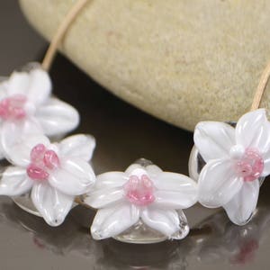 May include: A necklace made of white glass beads shaped like flowers with pink centers. The beads are strung on a clear cord.