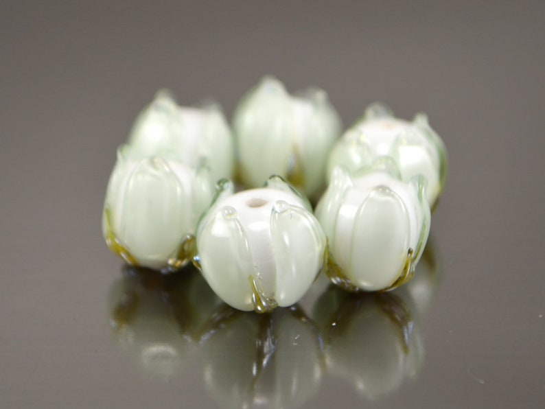 May include: A set of six white glass beads with green accents, shaped like flowers. The beads are arranged in a circle.