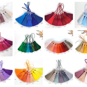Puede incluir: Una colección de 100 borlas de seda de diferentes colores, cada una con un lazo para colgar. Las borlas están dispuestas en filas de 10, con cada fila presentando un color diferente. Los colores incluyen tonos de rosa, azul, verde, rojo, naranja, amarillo, morado y marrón.