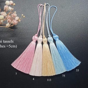 May include: Five mini tassels in pink, white, beige, light blue, and blue. The tassels are 2 inches long, or 5 cm. The tassels are hanging from a dark surface. The numbers 7, 4, 113, 75, and 33 are written below the tassels.
