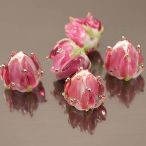 May include: Five pink glass flower beads with white centers and green accents. The beads are arranged in a cluster on a reflective surface.