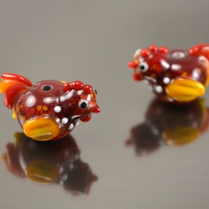 May include: Two red, yellow, and white glass chicken beads. The beads are speckled with small dots and have a hole through the center.